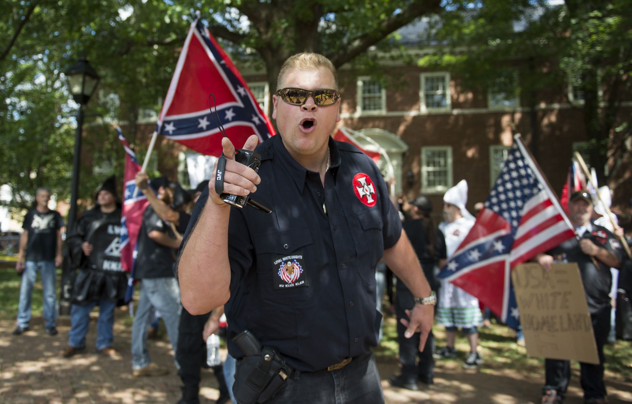 A large Caucasian man wearing a dark blue shirt with 'Loyal White Knights' and KKK patches shouts while pointing a finger. He wears sunglasses and has a handgun in a holster. Behind him, protesters hold Confederate and American flags in front of a brick building.