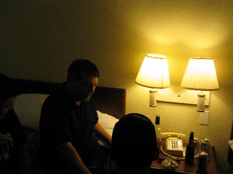 A dimly lit photograph of three men in a motel room. One man with dark hair and a striped shirt sits on the bed, while two others are partially visible in the foreground. A nightstand holds beer cans, a bottle, and a landline phone under the glow of two lamps.