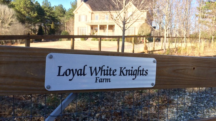 A photograph of a sign on a wooden fence that reads 'Loyal White Knights Farm' with a large house in the background.