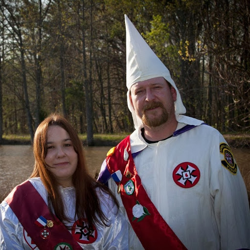 A photograph of a man and a woman in Ku Klux Klan regalia standing outdoors. The man wears a white robe, a pointed hood, and a red sash with medals. The woman wears a white robe and a red sash. They are positioned in front of a wooded area and a pond.