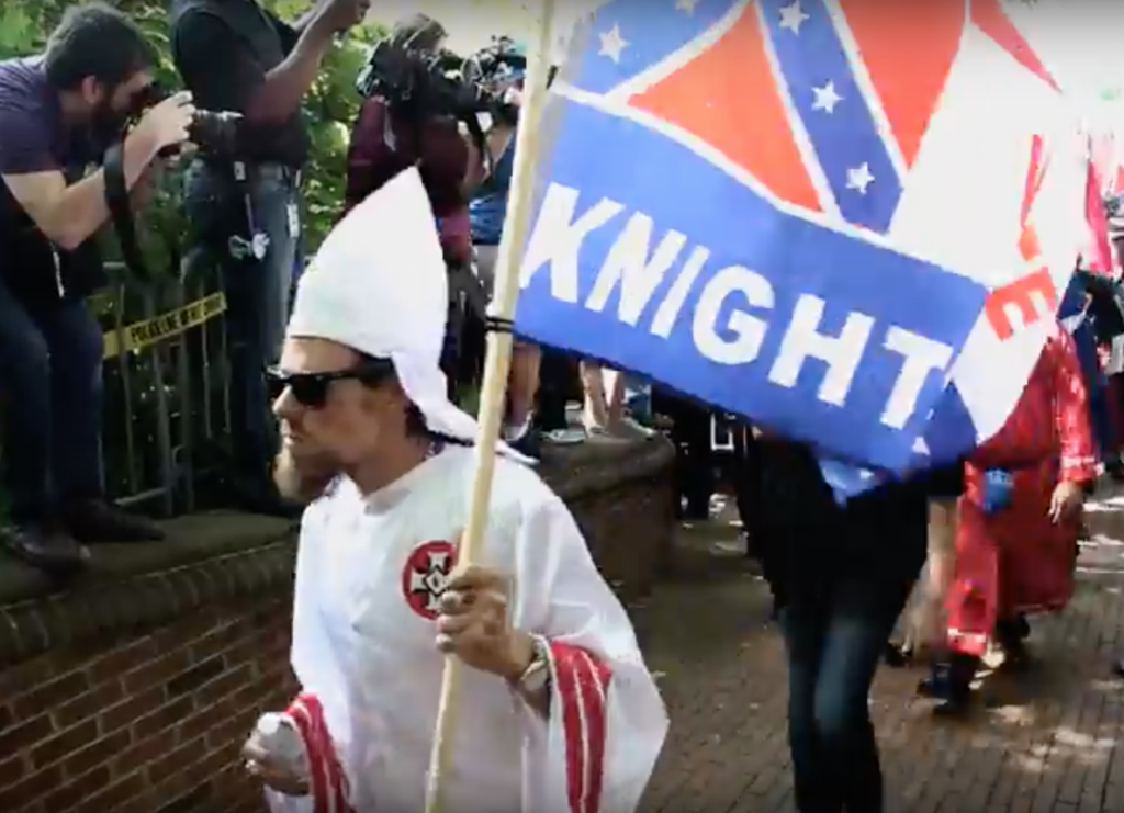 A photograph of a man in a white Ku Klux Klan robe and hood, wearing sunglasses and carrying a flag with the word 'KNIGHT' and Confederate imagery. A photographer is seen on the left capturing the scene.