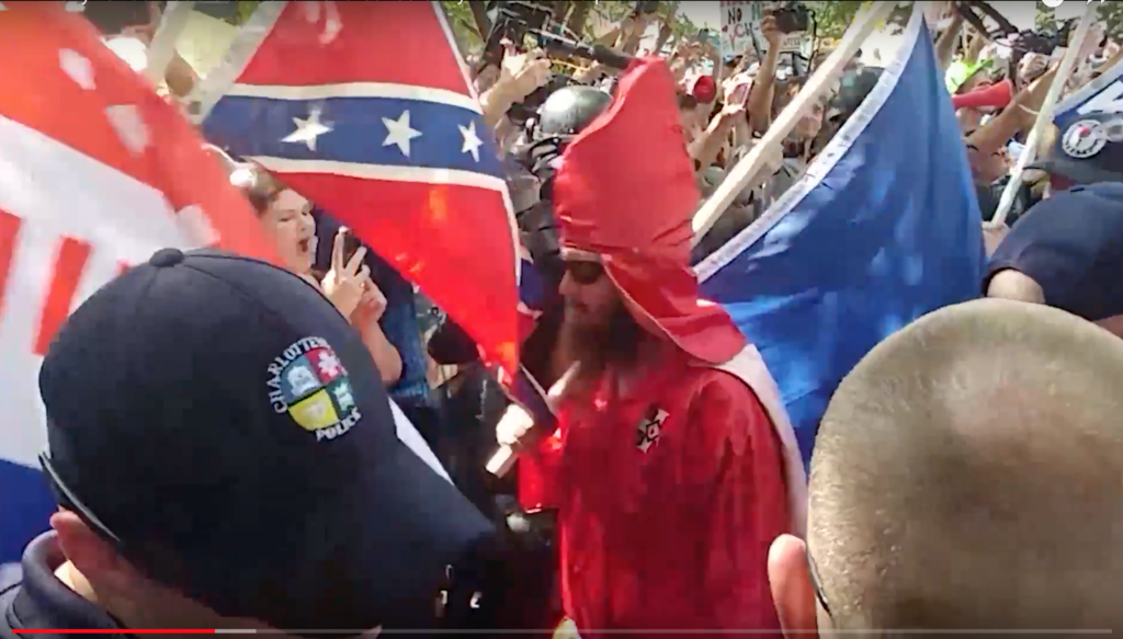 A photograph from a rally showing a man in a red KKK robe and hood holding a Confederate flag, surrounded by a crowd and a police officer.