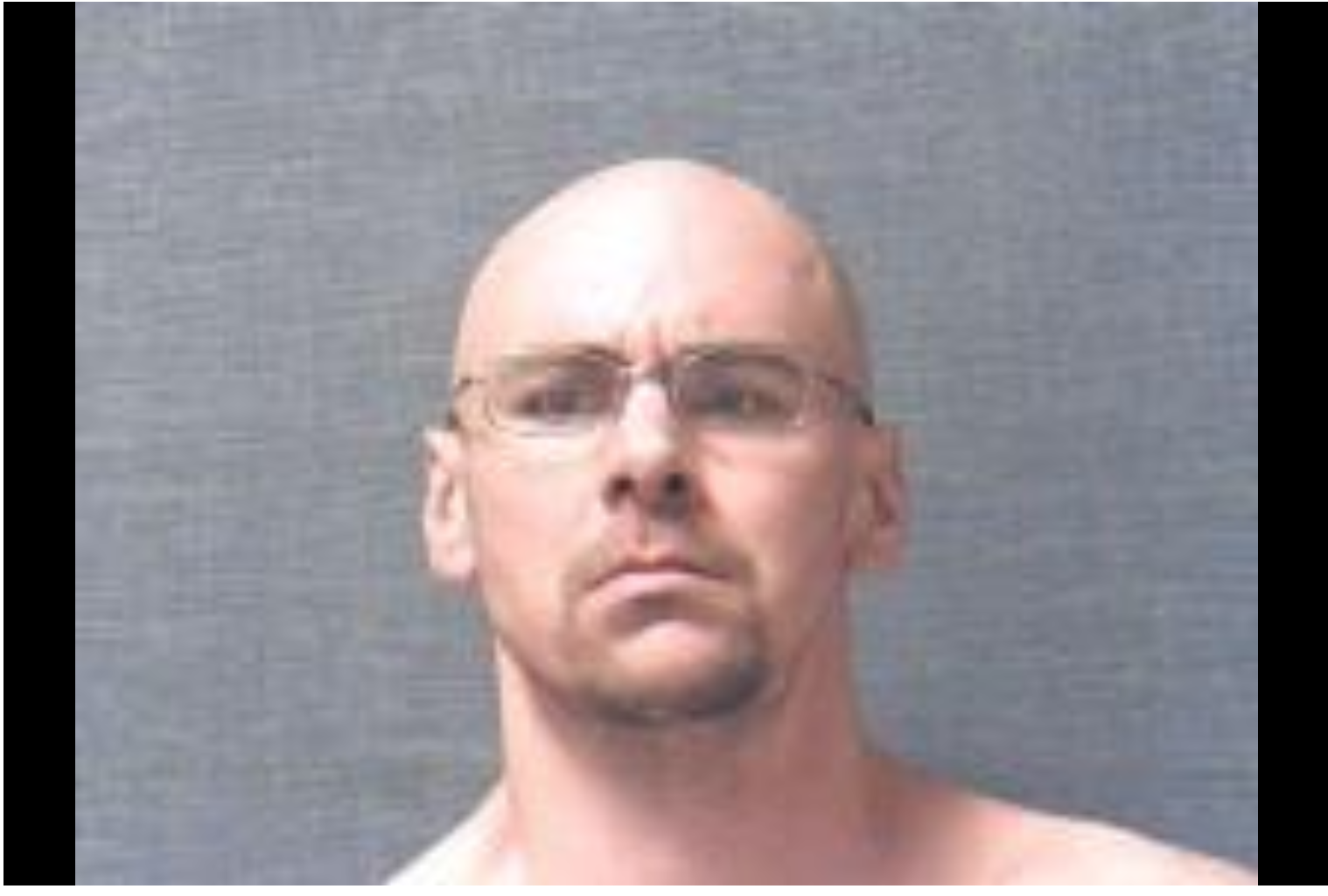 A mugshot of a bald Caucasian man with glasses and a goatee against a grey background.