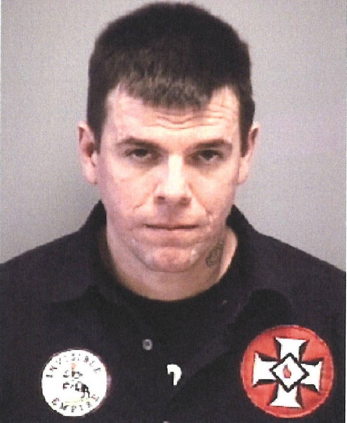 A mugshot-style photograph of a Caucasian man with short dark hair, wearing a black shirt with KKK-related patches, including a blood drop cross and a circular white patch.