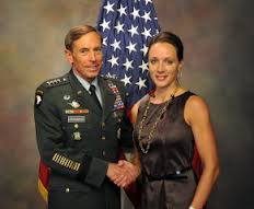 A formal photograph of General David Petraeus in a full military dress uniform shaking hands with Paula Broadwell, who is wearing a dark sleeveless dress, in front of an American flag.