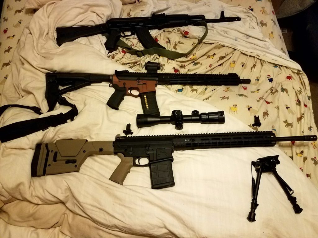Three rifles, including an AK-style and two AR-style rifles, along with a scope and a bipod, are laid out on a bed with dog-patterned sheets.
