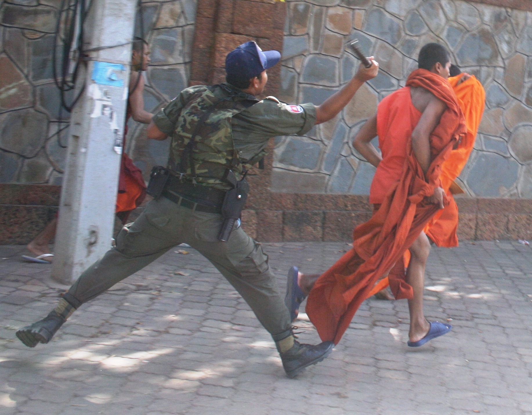 A Cambodian security officer in a camouflage vest and olive green uniform swings a baton at a Buddhist monk in orange robes who is fleeing on a paved street.