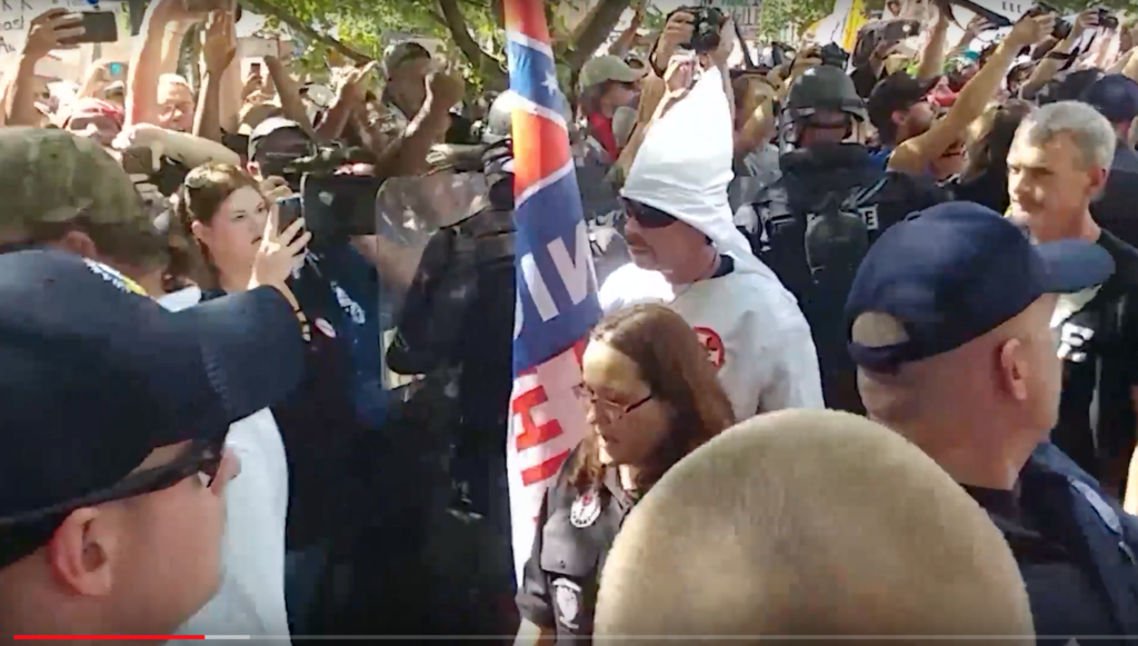 A video screenshot of a protest featuring a member of the KKK in a white hood and robe. The scene is crowded with police, protesters, and media. A Confederate flag is visible. In the foreground, a bald man is seen from the back.