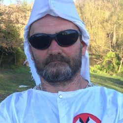 A close-up photograph of a Caucasian man wearing a white Ku Klux Klan robe and hood, dark sunglasses, and a full beard, standing in an outdoor setting.