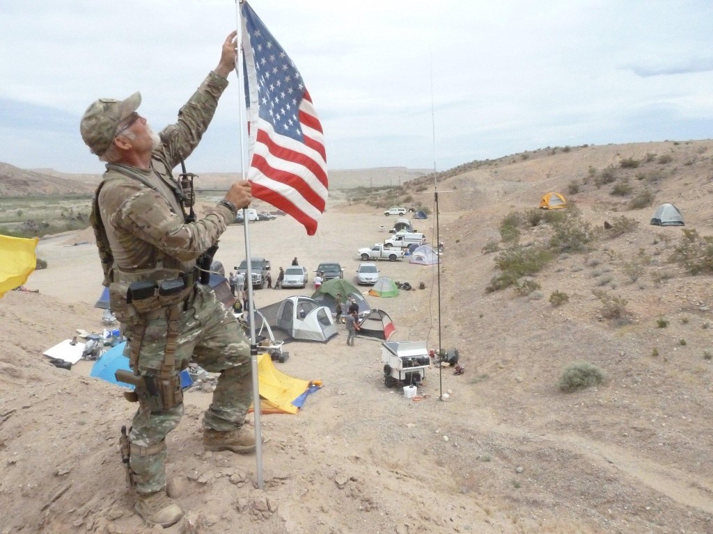 A man in full camouflage tactical gear stands on a desert ridge, holding a flagpole with the American flag. Below him is a campsite with several tents and parked vehicles in a dry, hilly landscape.