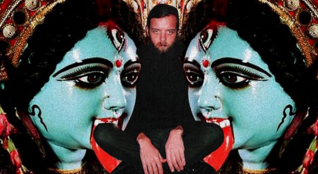 A man with dark hair and a beard sits cross-legged in front of a mirrored background featuring the blue-skinned Hindu goddess Kali.