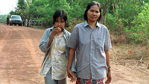 A woman and a young girl walking hand-in-hand on a dirt road in a rural area, likely in Cambodia. They are both wearing button-down shirts. In the background, a white SUV and a few figures are visible near a gate.