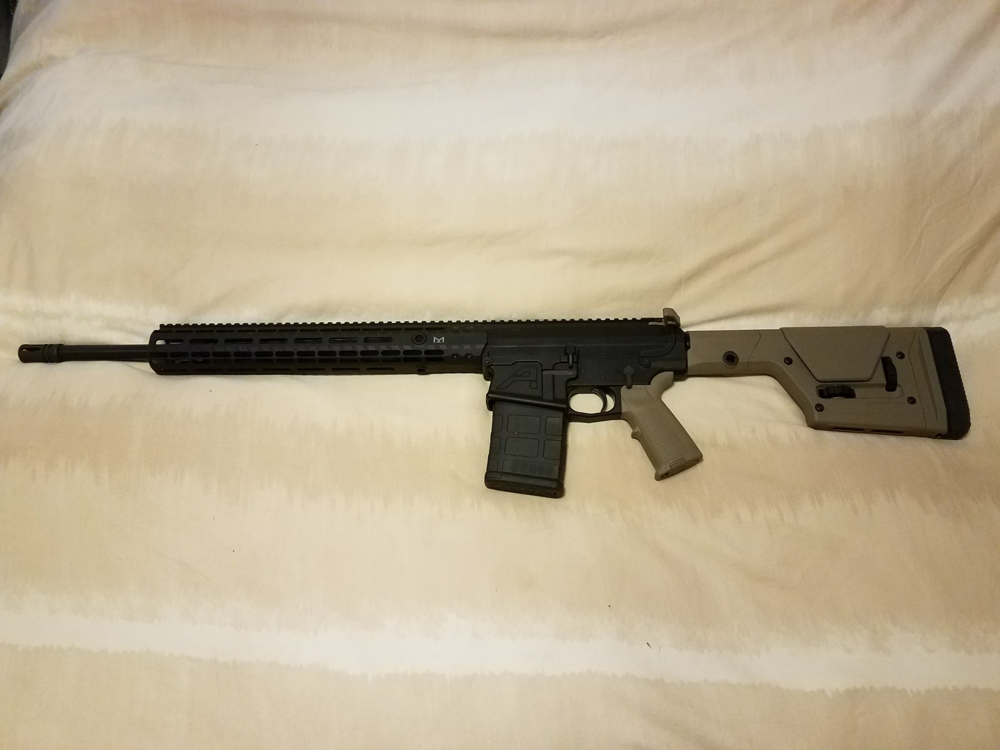 A photograph of a semi-automatic rifle with a black finish and tan-colored stock and grip, lying on a light-colored textured surface.
