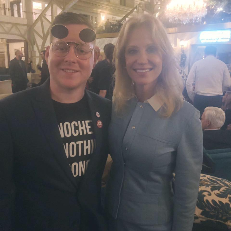 A photograph of Richard Spencer and Kellyanne Conway standing together at an indoor event. Spencer is wearing a black t-shirt under a blazer and glasses with flip-up lenses. Conway is wearing a light blue jacket.