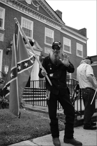 A black and white photograph of a man with a long, full beard and sunglasses, wearing a dark shirt and a baseball cap, holding a Confederate flag in front of a brick building that says 'COUNTY COURT HOUSE'. He is waving with his left hand.