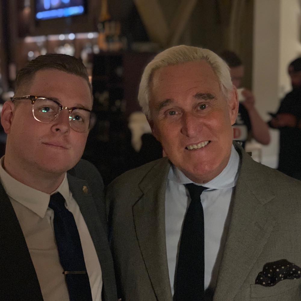 A photograph of two men in suits standing close together. On the right is Roger Stone, an older man with white hair. On the left is a younger man with short brown hair and glasses.