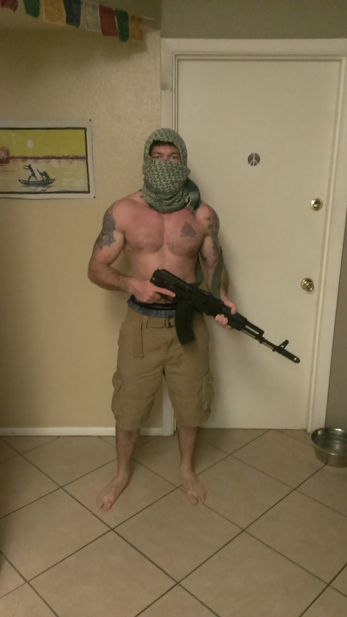 A shirtless Caucasian man with tattoos on his arms and chest, wearing a green and black keffiyeh over his face and tan cargo shorts, holding a black semi-automatic rifle in an indoor setting.