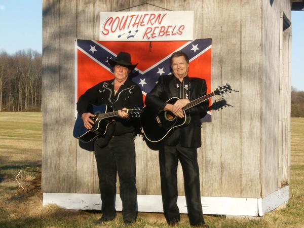 Two men holding acoustic guitars stand in front of a wooden shed adorned with a Confederate flag and a sign reading 'SOUTHERN REBELS'. One man wears a cowboy hat, while the other has a full head of hair and wears a suit jacket.