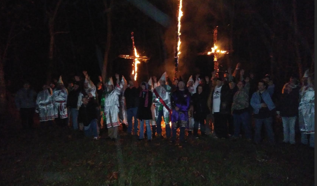 A nighttime photograph of a group of people, some in KKK robes and hoods, standing in front of three large burning crosses in a wooded area. Many individuals have their right arms raised in a salute.