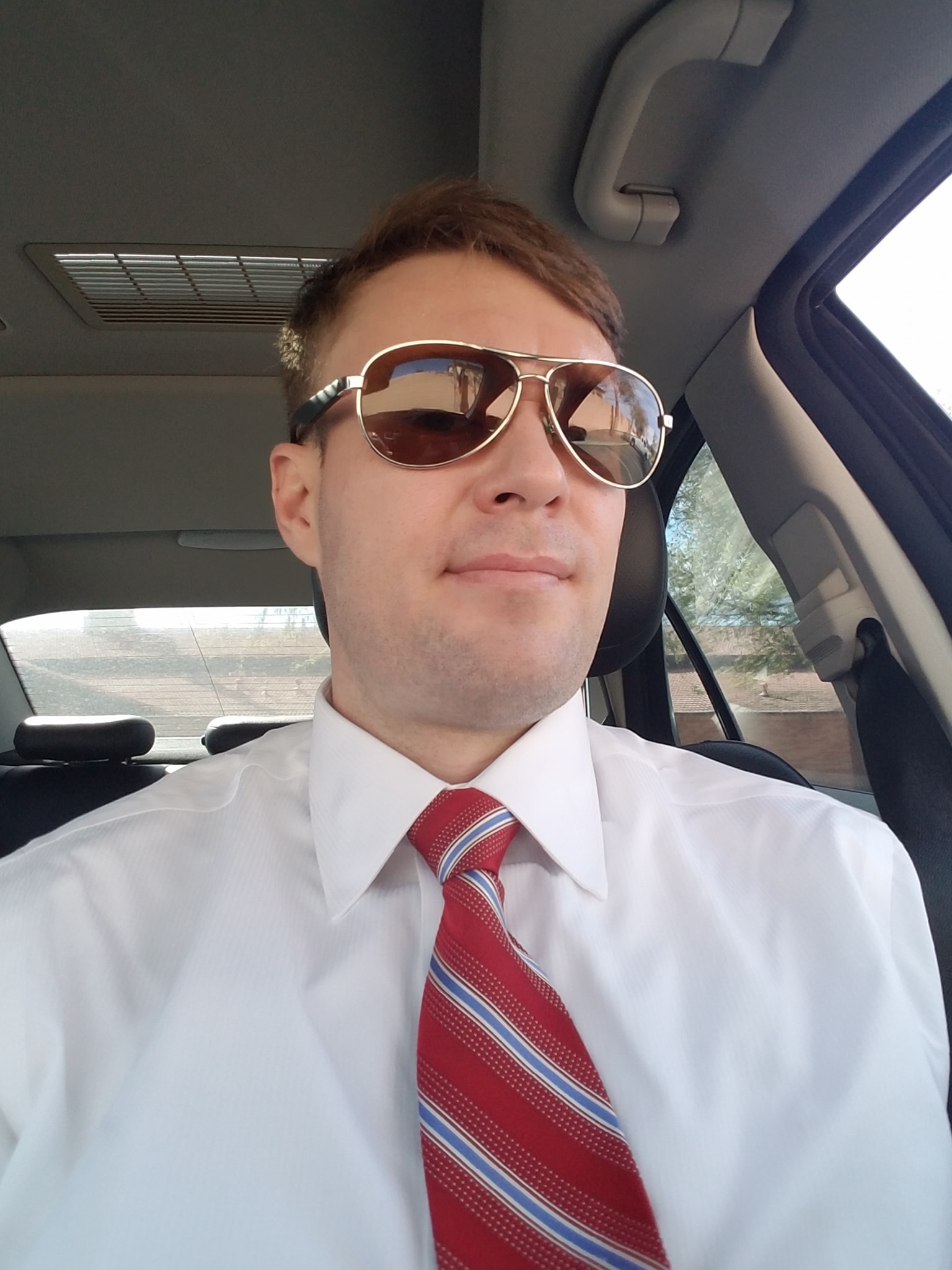 A selfie of a Caucasian man with short brown hair wearing aviator sunglasses, a white dress shirt, and a red striped tie, sitting inside a car.