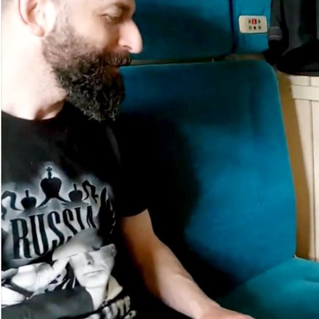 A side profile photograph of a man with a thick dark beard and short dark hair, wearing a black t-shirt with the word 'RUSSIA' and a graphic. He is seated in a blue upholstered chair.