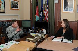 A photograph of General David Petraeus in military uniform sitting at a desk across from Paula Broadwell in an office with flags in the background.