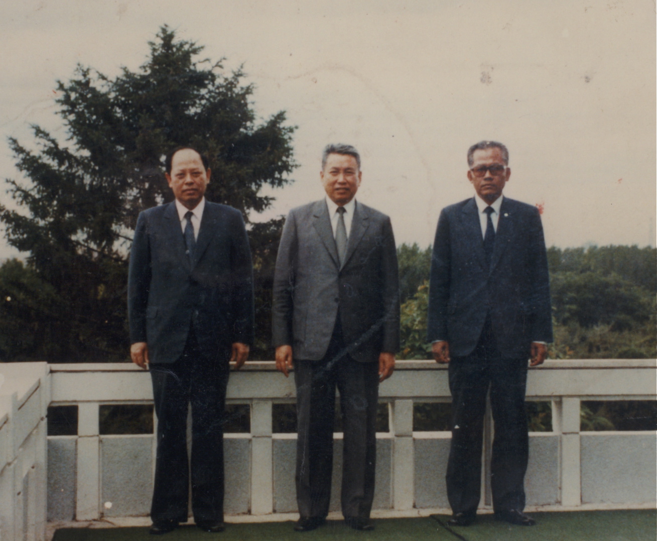 A historical photograph of three Asian men in suits, including Pol Pot in the center, standing on a balcony with a background of trees.