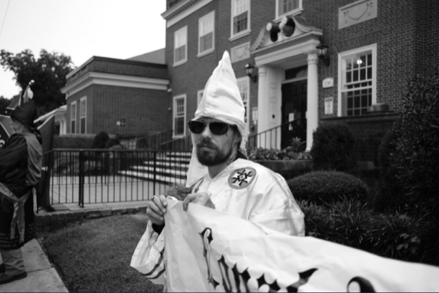 A black and white photograph of a man wearing a white KKK robe and pointed hood, sunglasses, and a full beard, holding a banner in front of a brick building.