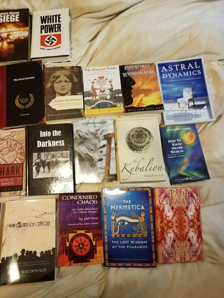 A collection of books laid out on a light-colored fabric surface. The titles include extremist literature such as 'Siege' by James Mason, 'White Power' by George Lincoln Rockwell, and 'The Culture of Critique' by Kevin MacDonald, alongside various occult and esoteric texts like 'The Kybalion', 'The Hermetica', and 'Condensed Chaos'.