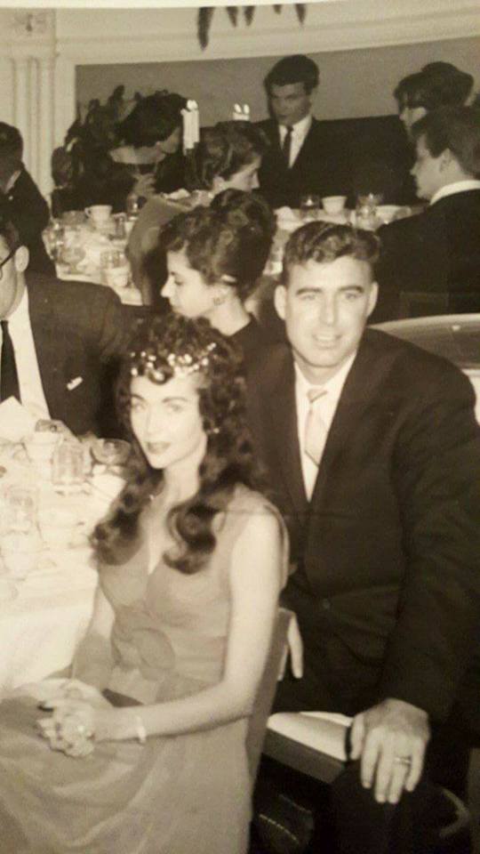 A vintage black and white photograph of a man and a woman seated at a formal dinner table. The man is wearing a dark suit and tie with a full head of hair, and the woman is wearing a sleeveless dress with long wavy hair. Other guests are visible in the background.