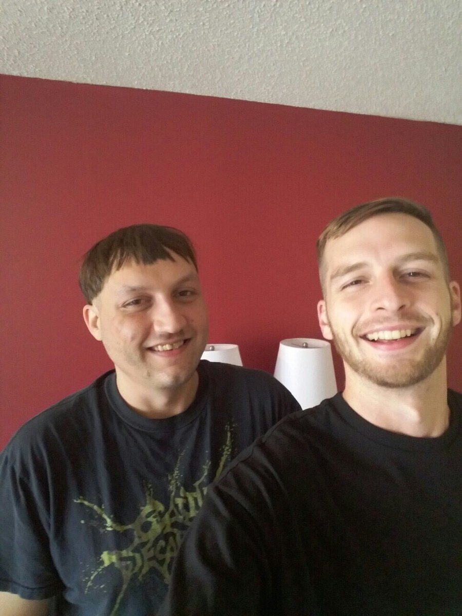 A selfie of two Caucasian men standing in front of a red wall. The man on the left has a bowl-cut hairstyle and is wearing a black graphic t-shirt. The man on the right is smiling, has short hair and a beard, and is wearing a black t-shirt.