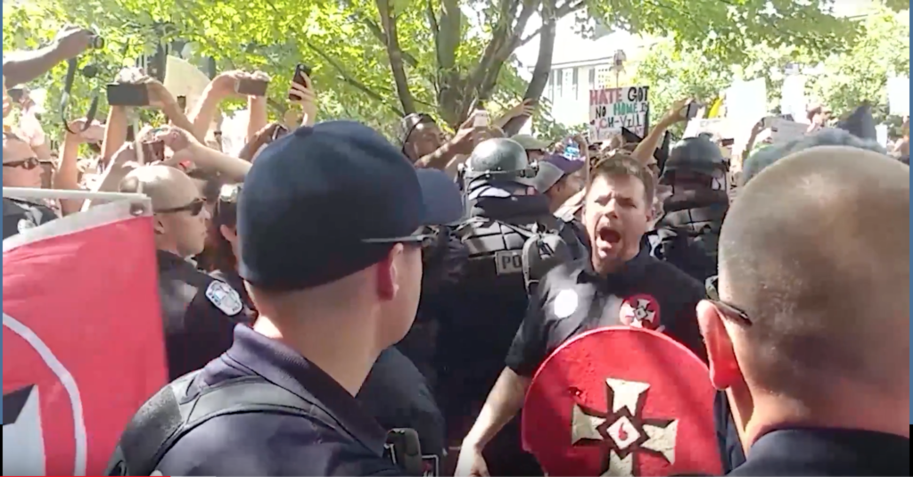A photograph from a white supremacist rally showing a man in a black shirt with a KKK patch shouting while holding a red shield. Police and other protesters are in the background.