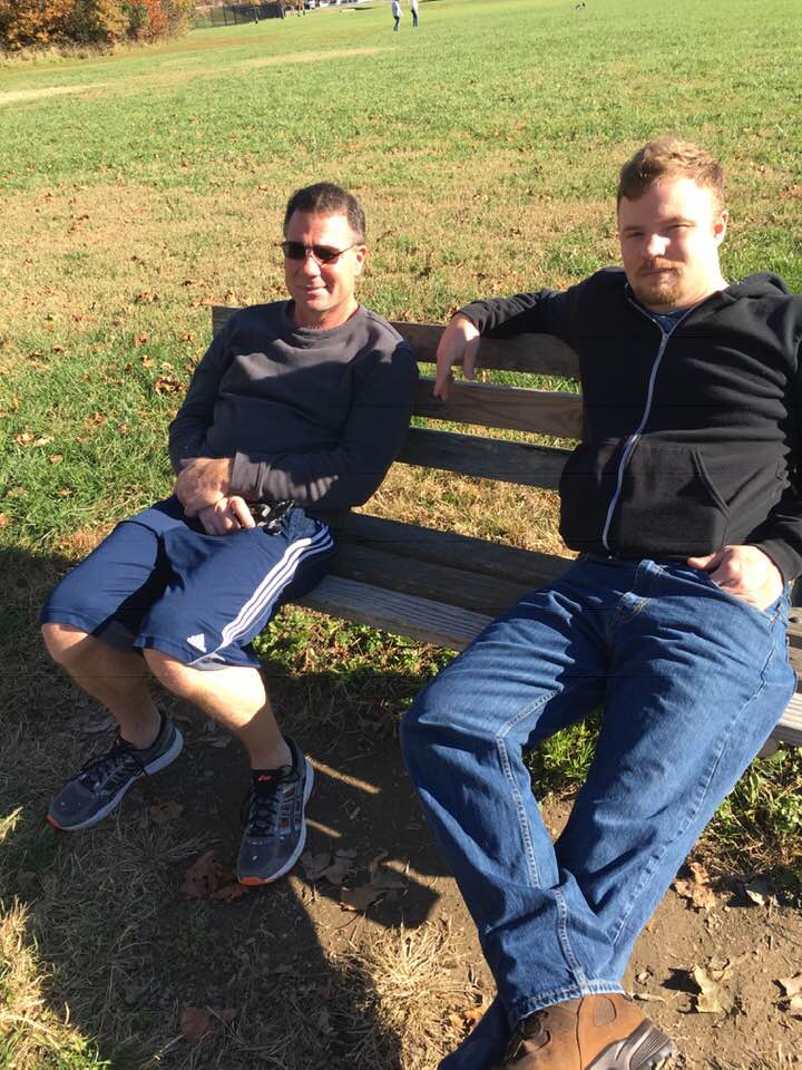 Two Caucasian men sitting on a wooden park bench in an open grassy field. The man on the left is wearing sunglasses, a grey long-sleeved shirt, and blue athletic shorts. The man on the right is wearing a black zip-up hoodie and blue jeans.