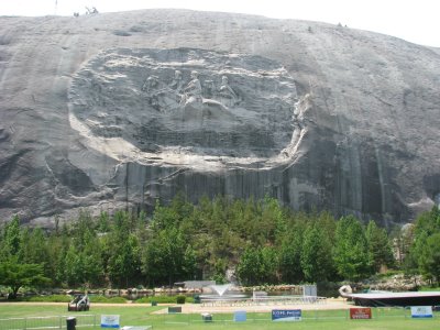 A wide-angle photograph of the Stone Mountain Confederate Memorial carving on a large granite rock face, with a park area and trees in the foreground.