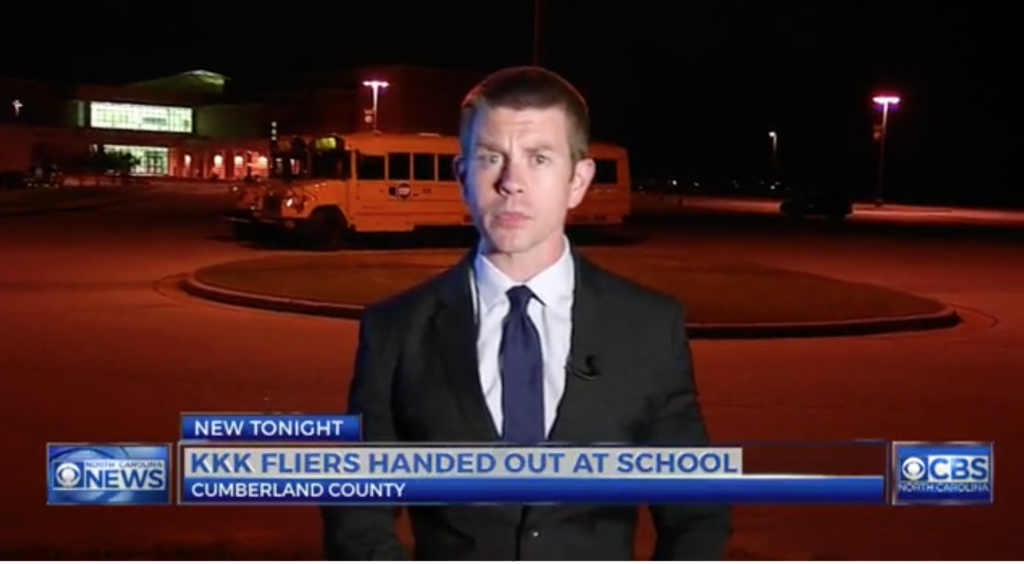 A news reporter stands in front of a school bus at night. A news graphic at the bottom reads 'KKK FLIERS HANDED OUT AT SCHOOL' in Cumberland County, North Carolina.
