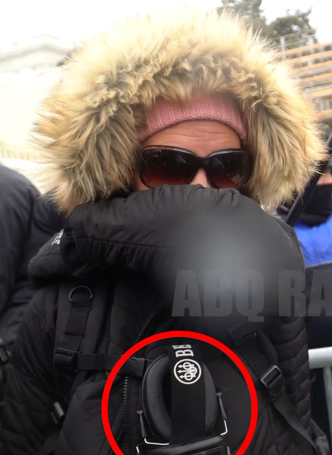 A close-up photograph of a person wearing a pink beanie, sunglasses, and a fur-lined hood, with their face partially obscured by their arm. A red circle highlights a pair of black earmuffs hanging from their black tactical-style jacket.