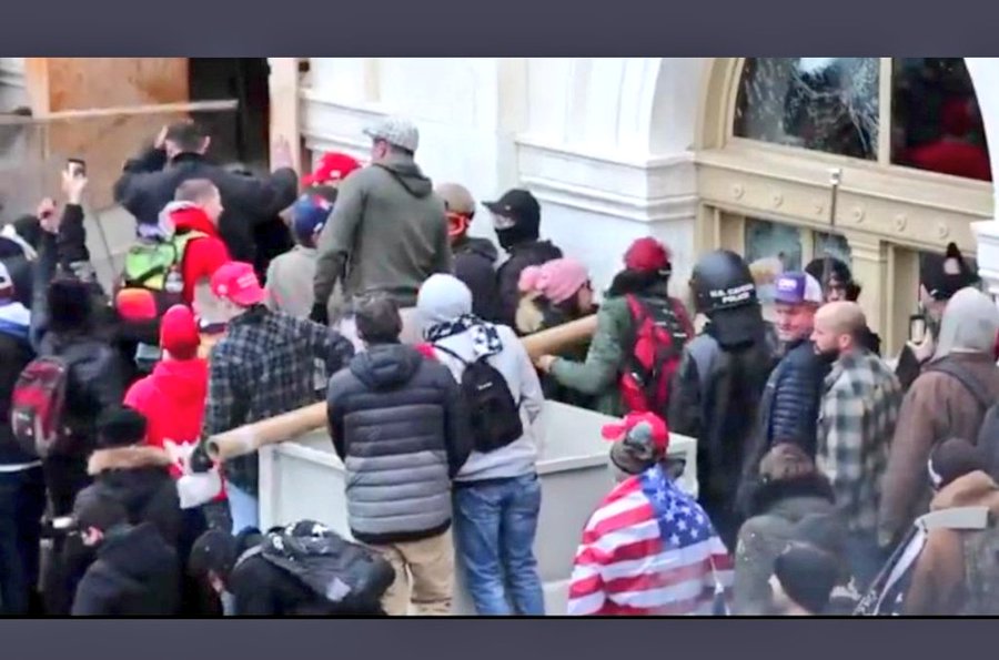 A photograph showing a crowd of rioters at the U.S. Capitol on January 6th, including a woman in a pink hat and several individuals in red hats, some carrying long objects near a broken window.