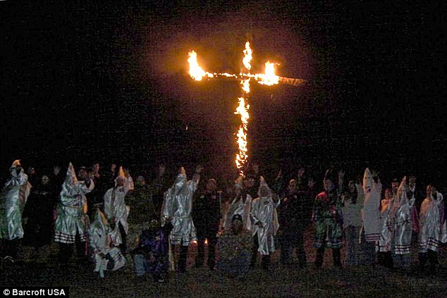 A group of individuals in Ku Klux Klan robes and hoods stand before a large burning cross at night.