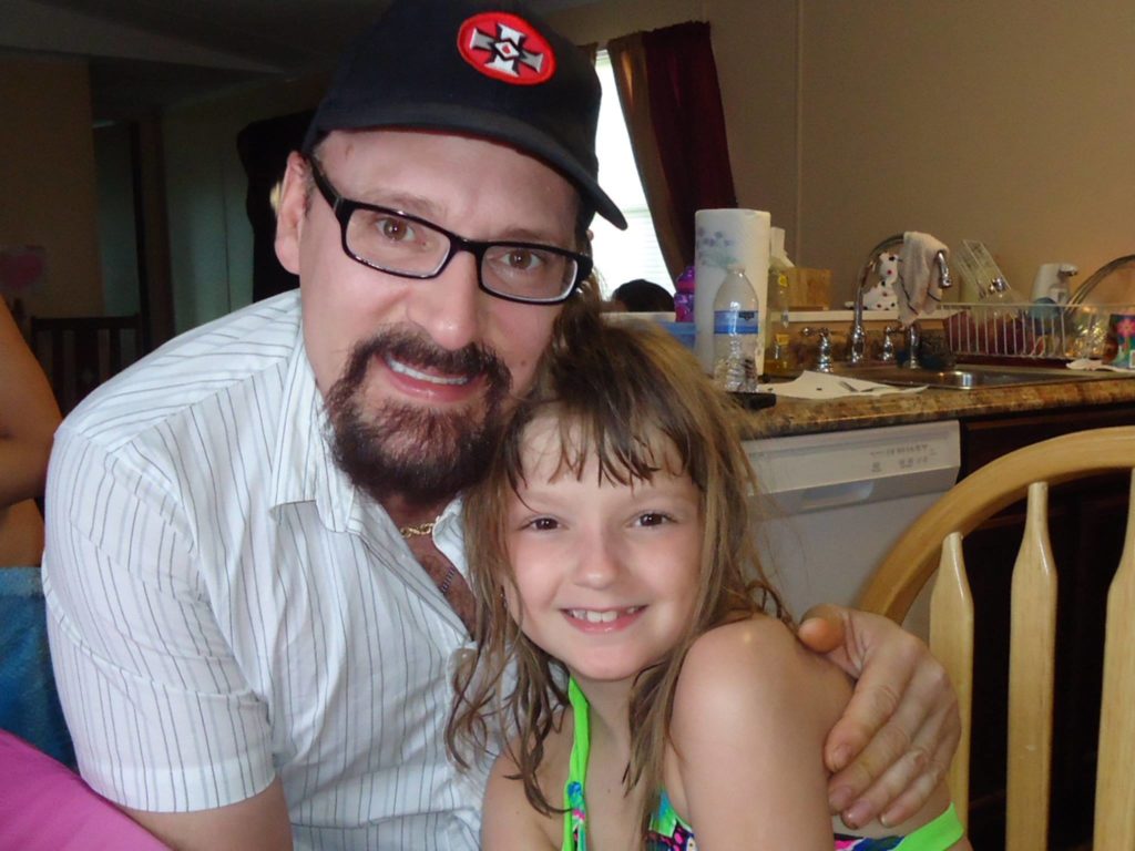 A photograph of a man wearing a black baseball cap with a KKK symbol, glasses, and a goatee, posing with a young girl in a domestic setting.
