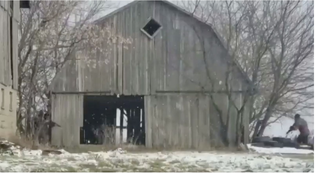 A weathered grey wooden barn in a snowy field with bare trees. A person in dark clothing is visible on the right side near some tires.