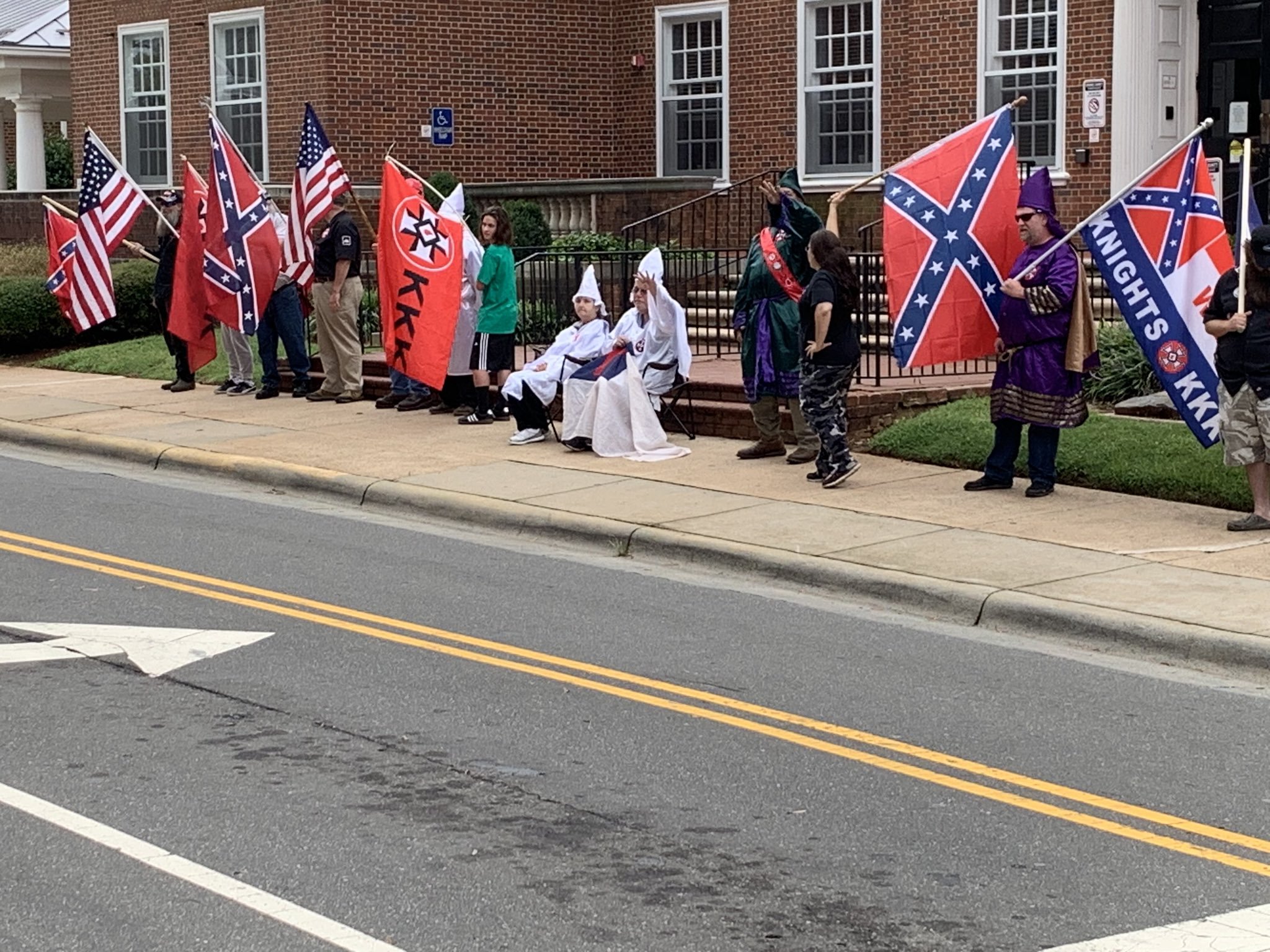 A photograph of a group of individuals participating in a KKK demonstration on a sidewalk in front of a brick building. They are holding American flags, Confederate flags, and a red flag with a KKK insignia. Several people are wearing traditional KKK robes and hoods in white, green, and purple.
