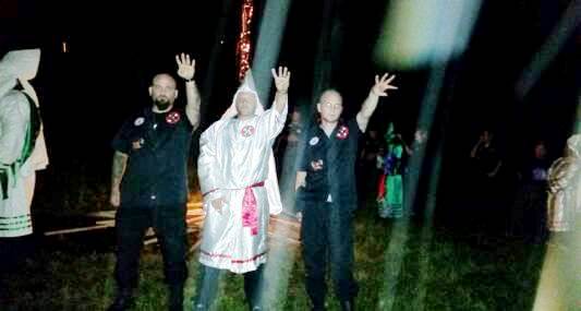 A nighttime photograph showing three men associated with the Ku Klux Klan. One man in the center wears a white robe and hood, while two men flanking him in black shirts perform a Nazi-style salute.