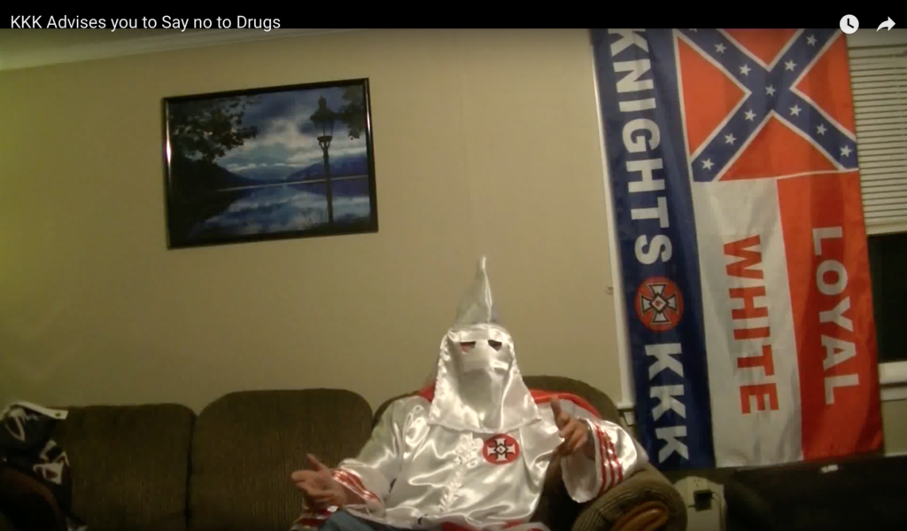 A screenshot of a video titled 'KKK Advises you to Say no to Drugs' showing a person in a white KKK robe and hood sitting on a couch next to a 'Loyal White Knights KKK' flag.