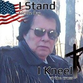 A portrait of Larry Mitchell Hopkins wearing sunglasses and a fringed jacket, with text overlays saying 'I Stand for the flag' and 'I Kneel at the cross' alongside an American flag and a cross.