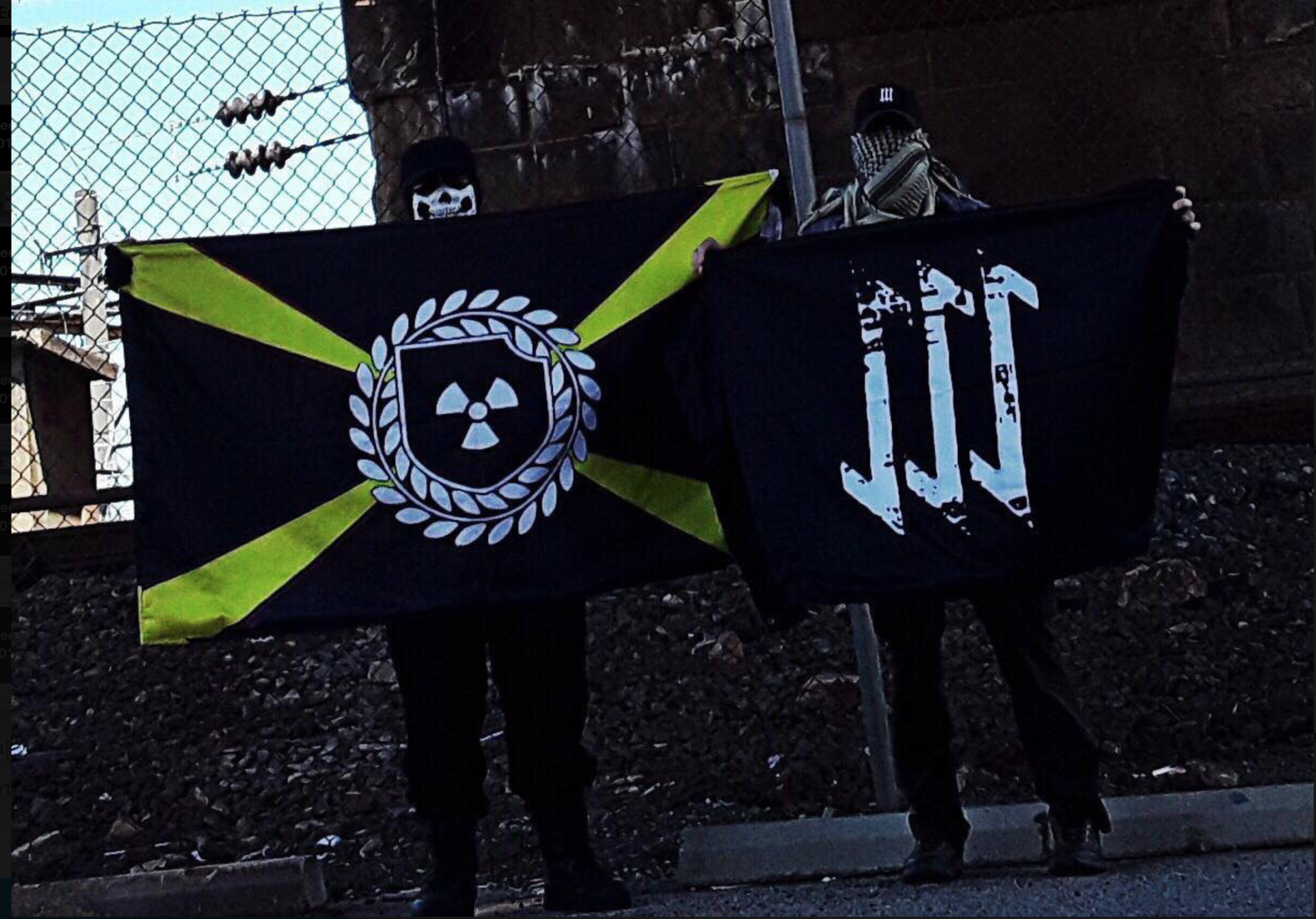Two masked individuals standing outdoors at night, holding flags associated with neo-Nazi extremist groups. One flag features a radiation symbol within a wreath on a black and yellow background, and the other features three stylized white symbols on a black background.