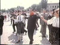 Kim Il Sung celebrating in the streets A historical photograph showing a man in a dark suit and glasses dancing in a circle with people wearing traditional folk costumes in an outdoor plaza.