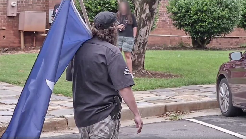 A man seen from behind wearing a black short-sleeved shirt with a patch, camouflage pants, and a black baseball cap over long curly hair. He is carrying a large blue flag over his shoulder. In the background, a person with a blurred face stands near a brick building and a tree.
