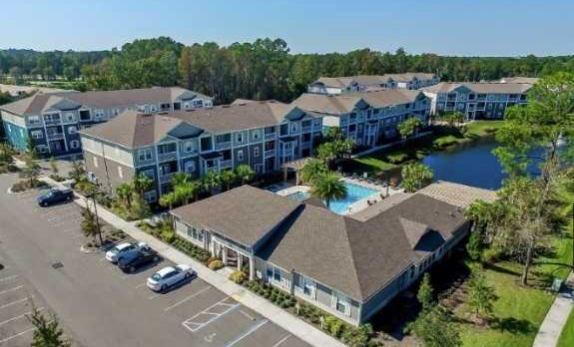 An aerial view of a modern apartment complex featuring several multi-story buildings, a swimming pool area, a parking lot with cars, and a small pond surrounded by greenery.