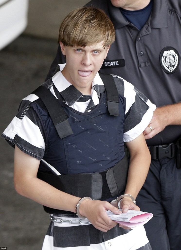 A photograph of Dylann Roof in a striped prison uniform and bulletproof vest, handcuffed and sticking his tongue out while being escorted by a police officer.