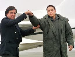 A photograph of two men standing in front of an airplane; one man in a dark suit is holding the arm of another man wearing a heavy green parka.